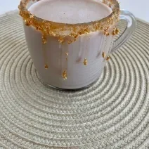 Fotografia em tons de marrom em uma bancada com um pano bege e uma xícara de vidro com a bebida de cappuccino de caramelo salgado dentro. Xícara decorada com calda de caramelo e biscoitos Tostines Especiarias