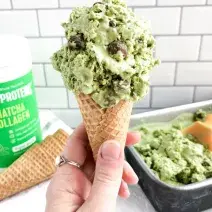 Fotografia em tons de verde em uma bancada de madeira, com um fundo de azulejo, com uma pessoa segurando uma casquinha com o sorvete de matcha, um pote de produto de Vital Proteins. E ao fundo, um recipiente com mais sorvete.
