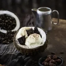 Fotografia de duas bolas de sorvete de coco com pedaços de chocolate dentro de um coco aberto, polvilhada com cacau em pó. O fruto está sobre uma mesa de madeira. Ao redor, cacau e nuts em potes pequenos na cor azul.