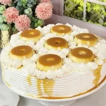 Foto da Receita de Bolo Recheado de Pudim com Doce de Leite Moça, decorado sobre um prato branco com sete mini pudins por cima, em uma bancada de mármore com flores ao fundo