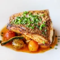 Fotografia de um filé de pescado ao papillote com alguns legumes ao forno por baixo do peixe. Por cima do peixe, que está em um prato branco, tem cebolinha salpicada.