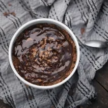 Fotografia vista de cima de um baked oats de air fryer dentro de um ramekin branco apoiado em um pano de tom cinza amassado, o qual está sobre uma madeira escura. Sobre o bolo tem uma calda de chocolate.