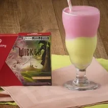 Foto aproximada de um copo alto com a receita de milkshake dividido em cores verde e rosa, servido sobre uma bancada de madeira com tecidos verde e rosa e com a caixa de KitKat Wicked ao lado esquerdo