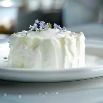 Fotografia em tons claros de um bolo de noiva com cobertura branca, sobre um apoio grande branco, apoiado sobre uma mesa de mármores branca. No bolo tem alguns enfeites florais roxos por cima.