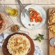 fotografia em tons de bege de uma bancada vista de cima, contém uma tábua redondo e marrom com uma pizza com fatias de banana, uma grade com uma pizza de chocolate e fatias de morango e um prato redondo e branco com uma fatia de pizza