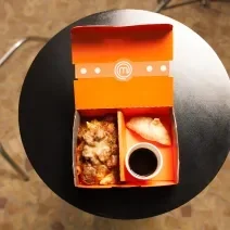 Fotografia em tons de marrom com uma mesa circular preta ao centro. Em cima da mesa existe um box com uma receita de ragu de costela e um mandu frito acompanhado com um molho escuro.