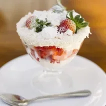Fotografia de uma taça de sobremesa com merengue, morangos, suspiro e granulado branco nas bordas. O recipiente está em cima de um prato branco de sobremesa, com uma colher pequena. O prato está sobre uma mesa marrom.