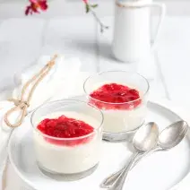 fotografia em tons de branco e vermelho tirada de um recipiente redondo branco, contém duas taças com mousse e calda de frutas vermelhas por cima