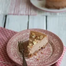 Fotografia de um prato de vidro desenhado com cor vermelho e branco. Sobre esse prato tem uma fatia de uma cheesecake de chocolate com amêndoas, ao lado de um garfo. Por cima da sobremesa tem lâminas de amêndoa. O prato está sobre um pano da mesma cor.