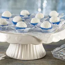 foto em tons de branco e azul de uma bancada cinza vista de frente, contém um suporte para servir bolos branco com diversos docinhos de coco com forminhas azuis