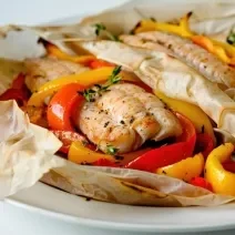 Fotografia de um filé de peixe no papillote com legumes por baixo cortados em fatias. Na fotografia são dois papillotes de peixe com legumes, um do lado do outro sobre um prato branco de vidro.