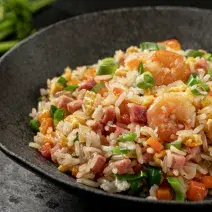 Foto aproximada de um prato preto fundo com a receita de arroz chop suey dentro, decorado com cebolinhas e com camarões e presuntos em cubos.