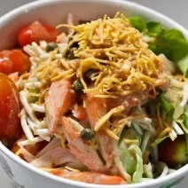 Fotografia de um recipiente branco fundo, e dentro, uma salada com batata palha, acelga, kani kama, agrião e tomates cereja. O recipiente está sobre uma mesa branca.