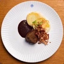 Fotografia vista de cima de um prato branco raso com molho de vinho tinto, cebolas fritas, filé mignon e batatas gratinadas. O prato está sobre uma bancada de madeira clara.