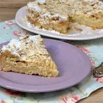 Fotografia em tons de lilás com um prato lilás ao centro. Em cima do prato existe uma receita de bolo de cor caramelo feito com farinha de amêndoas torradas.