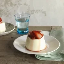 Fotografia de um flan de canela com calda de morangos em um prato branco fundo sobre uma mesa de madeira. Ao fundo, outro flan de canela e, ao lado, um copo d'água.