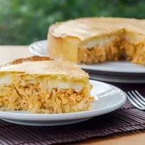 Fotografia de uma fatia de torta de frango com requeijão sobre um prato pequeno de vidro raso. No fundo tem um apoio branco e preto com o restante da torta quase inteira.