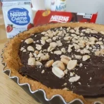 Fotografia de uma torta inteira charge ainda dentro da forma de alumínio. Por cima da torta tem amendoim, no fundo tem creme de leite e chocolate. A forma está sobre uma tábua de madeira.