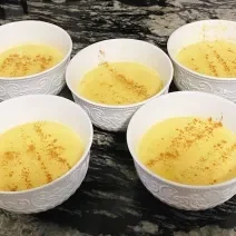 Foto de cinco potes brancos com a receita de curau cremoso em tom amarelo, polvilhado com canela em pó