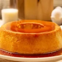 Fotografia mostra uma torta com textura delicada e cremosa, coberta por uma calda de caramelo dourada e servida em um prato branco.