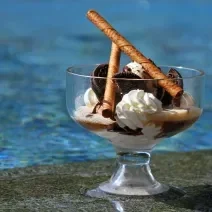 Fotografia de uma taça grande decorativa com cassata de chantilly e sorvete de Chocolate com alguns biscoitos e dois tubinhos de chocolate. O recipiente está apoiado sobre a base de uma piscina.