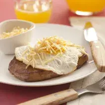 fotografia em tons de rosa, branco e marrom de uma bancada rosa vista de frente, contém um prato redondo branco com um pedaço de carne com ovo por cima e batata palha. Ao lado garfo e faca para servir, e ao fundo uma jarra e um copo transparente com suco.