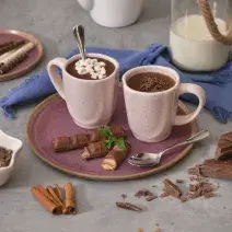 Foto de duas canecas em tom rosa claro, contendo a receita de Chocolate Quente, sobre um prato em tom violeta, com um tecido em tom azul atrás, uma jarra com leite e pedaços de chocolate e canelas em pau sobre a mesa