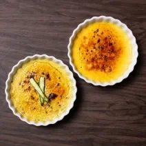 Fotografia vista de cima de dois ramekins brancos, um com creme brulée de baunilha com laranja, e o outro creme brulée de aspargos com bacon. Os ramekins estão sobre uma mesa de madeira.