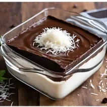 Fotografia de um recipinete quadrado de vidro com creme de coco e por cima, uma ganache de chocolate. Tem coco ralado decorando o chocolate, o recipinete está sobre uma mesa de madeira.