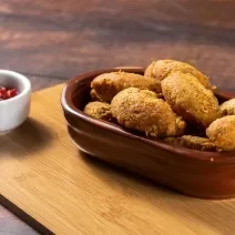 Fotografia de uma travessa marrom com vários bolinhos fritos em tons dourados por cima de uma tábua de madeira. Ao lado, um pequeno recipiente branco com um molho de pimentas vermelho.