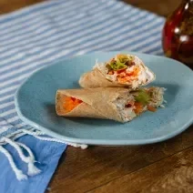 Fotografia em tons de azul com um prato azul ao centro. Em cima do prato existe uma receite de wrap recheada com frango desfiado, creme de ricota, tomate e alface.