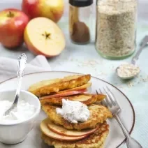 Fotografia de fatias de maçã com creme de leite, canela e aveia em um prato branco fundo, com um recipiente pequeno ao lado cheio de creme de leite. Ao fundo tem duas maçãs inteiras e uma metade, também tem canela.