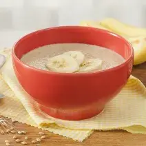 Fotografia em tons de amarelo e vermelho de uma bancada de madeira com um paninho amarelo e um recipiente redondo vermelho, sobre ele o mingau de farinha láctea com fatias de banana. Ao fundo duas bananas e aveias espalhadas pela bancada.