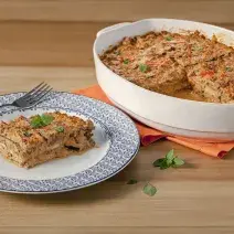 Fotografia em tons de azul e laranja em uma bancada de madeira clara, um pano laranja, um prato redondo branco com detalhes azuis e uma fatia da lasanha de vegetais com molho à bolonhesa em cima dele. Ao lado, um recipiente branco oval com a lasanha.