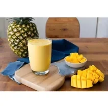 Fotografia de um copo de vidro com smoothie de iogurte, manga e abacaxi. O copo está sobre uma mini tábua de madeira, ao lado de dois recipientes brancos pequenos com pedaços de manga. No fundo tem um abacaxi inteiro sobre um pano azul escuro.