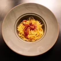 Fotografia em tons de amarelo com um prato cinza ao centro. Dentro do prato existe uma receita de macarrão carbonara finalizada com pedaços de guanciale frito.