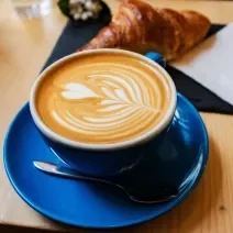 Foto da receita de Flat White. Observa-se uma xícara azul sobre um pires da mesma cor e a bebida dentro, com um formato de coração.