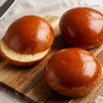 Fotografia em tons de marrom com uma tábua de madeira ao centro. Em cima da tábua existe três pãezinhos dourados por fora e branquinhos por dentro.