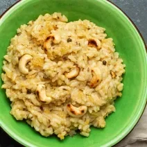 Fotografia vista de cima de um arroz crocante com castanha de caju dentro de um recipiente fundo verde, o qual está sobre uma mesa escura preta. Ao lado do recipiente tem um paninho e talheres por cima.