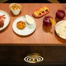 Fotografia de uma mesa de madeira com um brunch com pão sírio, hommus, ovos, frutas, massa folhada, creme inglês de requeijão, crumble de banana e torrada. Todas as partes estão porcionadas em recipientes brancos e separados.