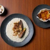 Fotografia vista de cima de três pratos pratos pretos com receitas diferentes, sendo elas: Fonduta de Gruyère com Cogumelos, Risotto de Brie com Abobrinha e Torta de Queijo com Compota de Frutas Amarelas. Os prato estão sobre uma bancada de madeira.