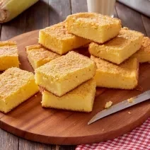 Foto da receita de bolo de milho cremoso, fatiado em pedaços sobre uma tábua de madeira sobre um pano quadriculado vermelho e branco. Na mesa há uma faca e espigas de milho