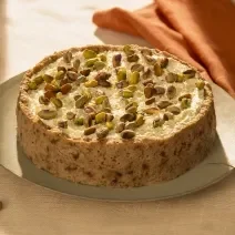 Foto da receita de torta de pistache com aveia corte rústico servida em uma porção grande sobre uma base de cerâmica em cima de uma mesa decorada com linho e um paninho laranja