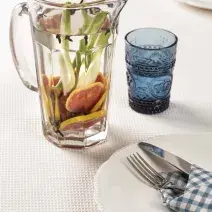 Fotografia em tons de azul em uma bancada branca com uma toalha branca, um guardanapo de pano azul xadrez. Ao centro, uma jarra de vidro com a água aromatizada e ao lado um copo de vidro azul com a água dentro.