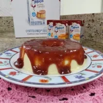 Imagem de um Pudim Romeu e Julieta sobre um prato colorido, numa bancada de cozinha com algumas embalagens do produto Moça Pra Toda a Família ao fundo