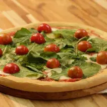 Fotografia em tons de dourado, verde e vermelho, ao centro pizza com folhas verdes e tomate-cereja sobre tábua redonda e guardanapo branco e vermelho, com talher ao lado, tudo sobre bancada de madeira em tons marrom.