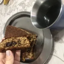 Fotografia de uma mão feminina segurando um pedaço de bolo de banana com aveia e chocolate. A outra parte do bolo está atrás da mão, sobre um recipiente oval de alumínio, ao lado de um copo de aluminio com café.