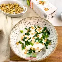 Foto vista de cima de uma mesa com a receita de creme de alho poró com inhame, num prato de cor clara, sobre uma tábua de madeira. Na mesa há potes com croutons, ervas e uma embalagem de bebida vegetal Nature's Heart