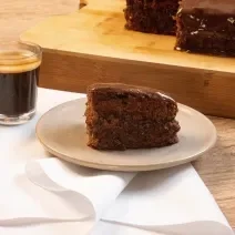 Fotografia de uma bancada de madeira, e sobre ela tem uma torta de chocolate com café em um prato de vidro em tom pastel, que está apoiado sobre um pano branco. Ao lado da sobremesa tem uma xícara com Nescafé, também sobre o pano.