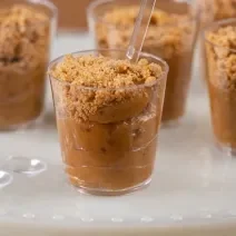 Fotografia em tons de branco com vários potinhos de plástico ao centro. Dentro de cada potinho existe um brigadeiro de cor caramelo feito com paçoca e coberto com paçoca triturada.